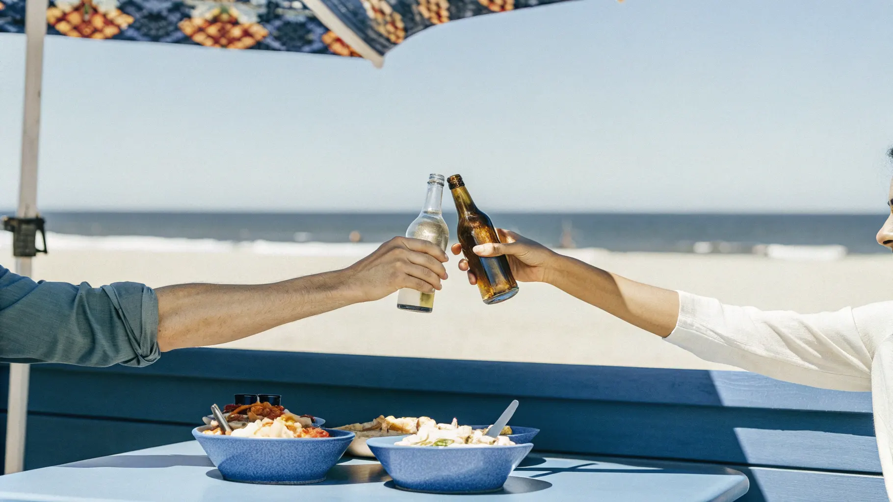 Friends toasting with beer bottles over shared plates of poke bowls, fish tacos, and nachos at a beachside restaurant